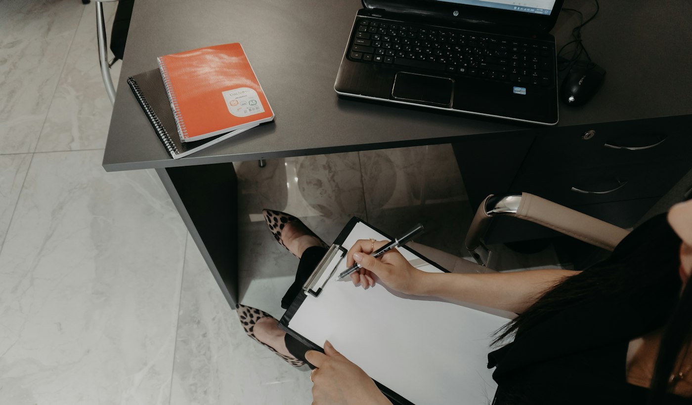 A person writing on a clipboard at a desk beside a laptop