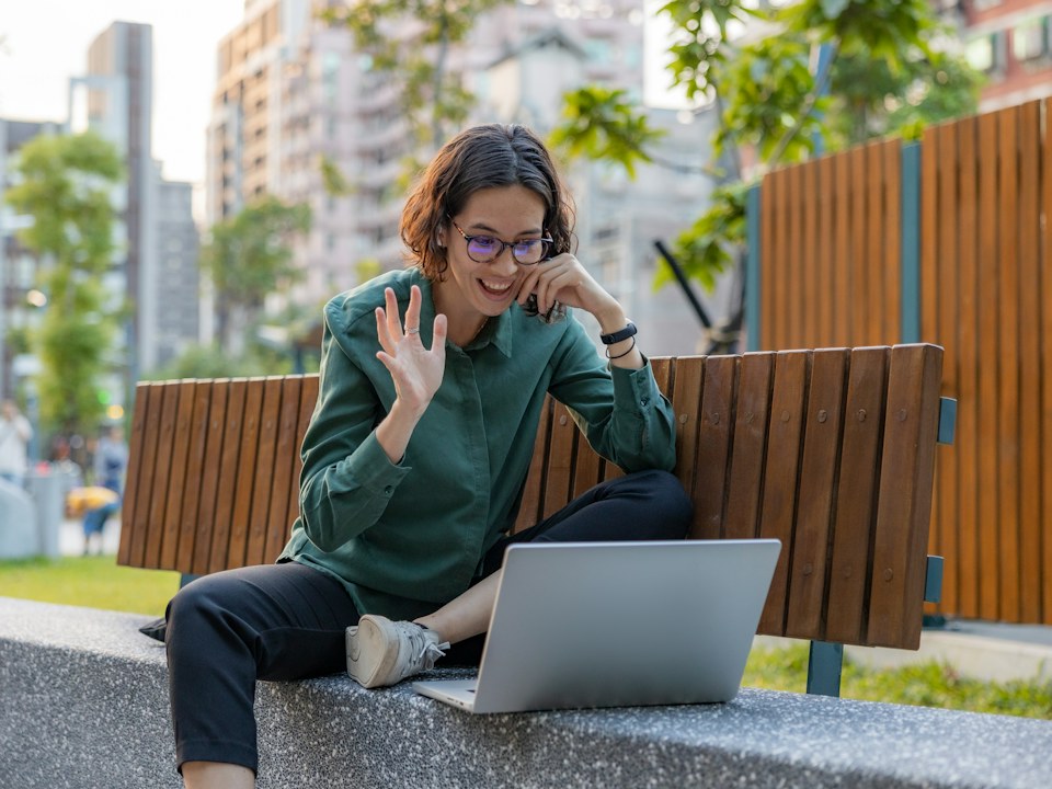 A woman on a video call with a laptop, sitting on a bench outside