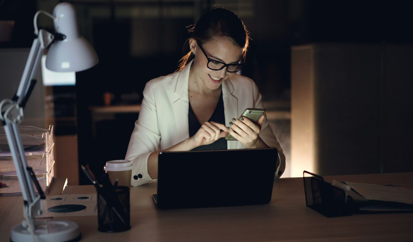 A woman working on a phone and a laptop at a desk late at night